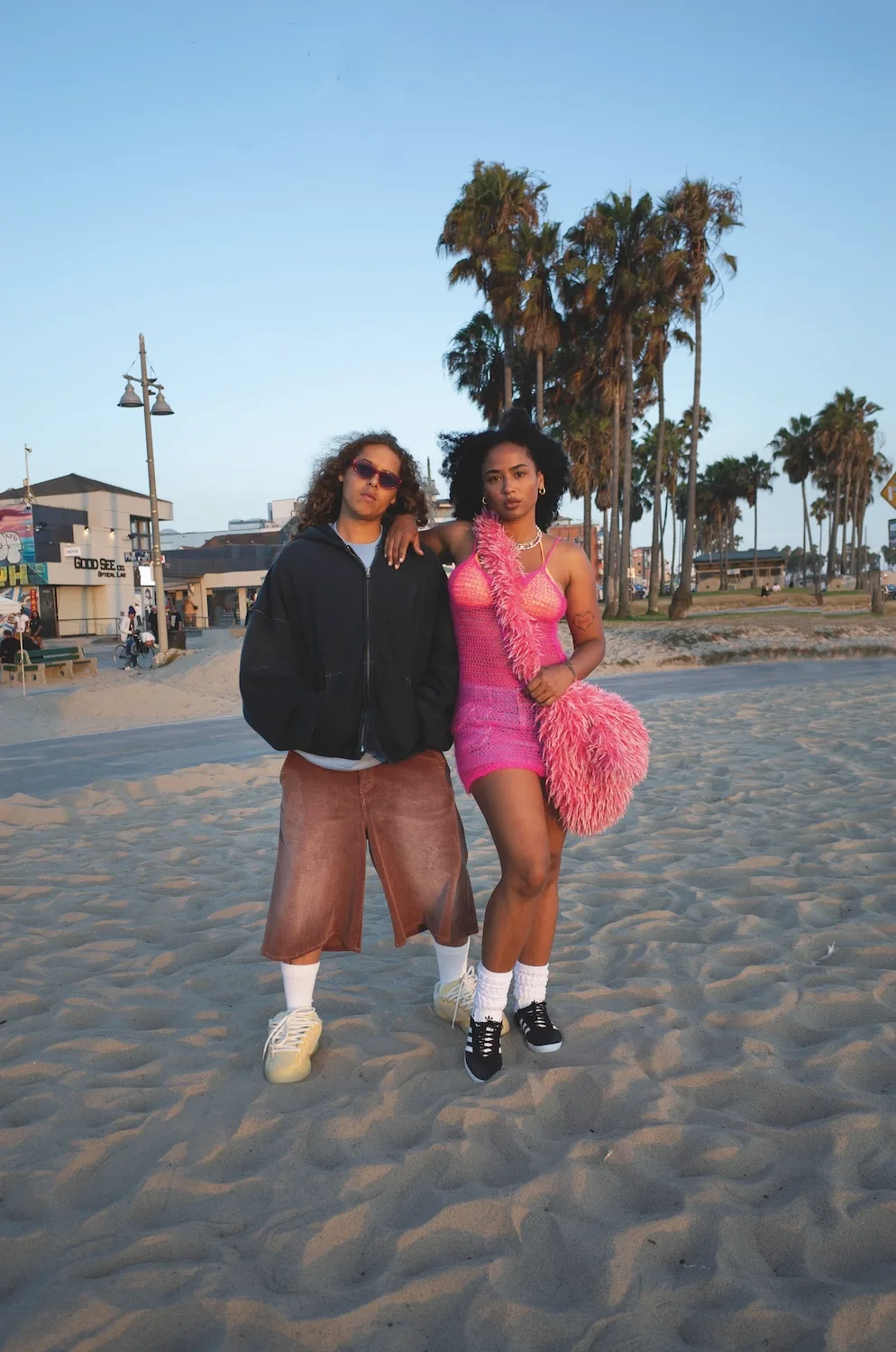 The cover photo of City of Angels: two people on the sand by a palm-lined boardwalk at golden hour, one in a black hoodie and long brown shorts, the other in a pink crochet dress with a pink feather boa