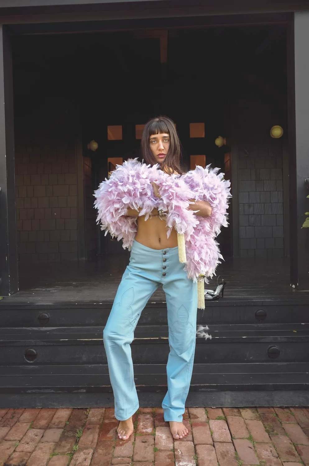 Jesse Jo Stark barefoot on brick paving in a lilac feather boa and pale blue leather trousers, in front of dark wooden steps and an open doorway
